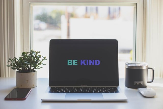 A laptop standing on a desk with the words 'be kind' written on the screen. Behind the computer there is a bright window.